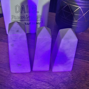 May include: Three white triangular-shaped crystal prisms, with a slightly iridescent sheen, are arranged in a row on a wooden surface. The prisms are illuminated with a purple light, creating a vibrant and ethereal effect.