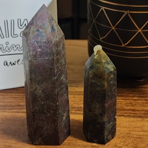 May include: Two dark purple and green crystal points, one larger than the other, with a pointed top and a flat base. The crystals are on a wooden surface.