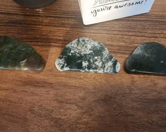 May include: Three cloud-shaped, green and white moss agate stones are displayed on a wooden surface. The stones vary in color and pattern, with one having a more translucent appearance. A small card with the text "you're awesome!" is in the background.