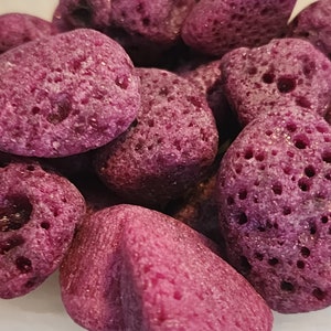 May include: Close-up of a collection of porous, oval-shaped stones in a deep purple hue. The stones have a rough texture with numerous small holes, creating a unique appearance. The stones are arranged in a white bowl.