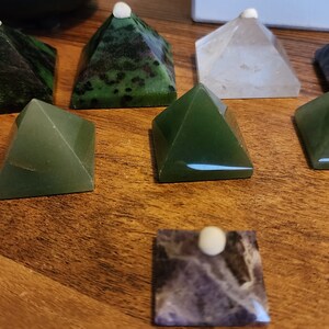 May include: Nine crystal pyramids, some green, some purple, and one clear, arranged on a wooden surface. Each pyramid has a small white dot on top.