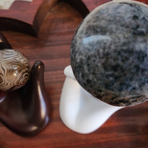 May include: Two polished spheres, one black and gray with a swirling pattern, and one white and brown with a swirling pattern, are displayed on a wooden surface. The larger sphere is on a white stand.