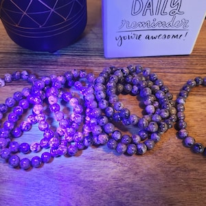 May include: A close-up of several purple and grey gemstone bracelets under a UV light. The bracelets are made of round beads and are stacked on top of each other.