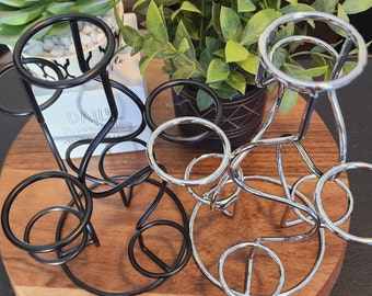 May include: Two metal drink holders, one black and one silver, with multiple circular openings for holding glasses or cups. The holders are on a wooden surface.