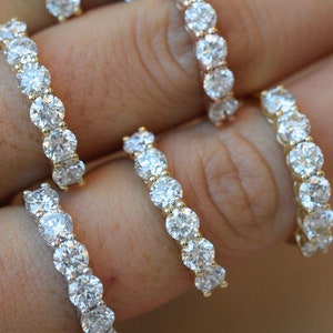 May include: Five gold rings with a row of clear round stones. The rings are stacked on a hand with brown nail polish.