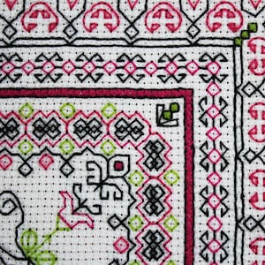May include: Close-up of a white fabric with a black, pink, and green embroidered design. The design features geometric shapes and floral motifs.