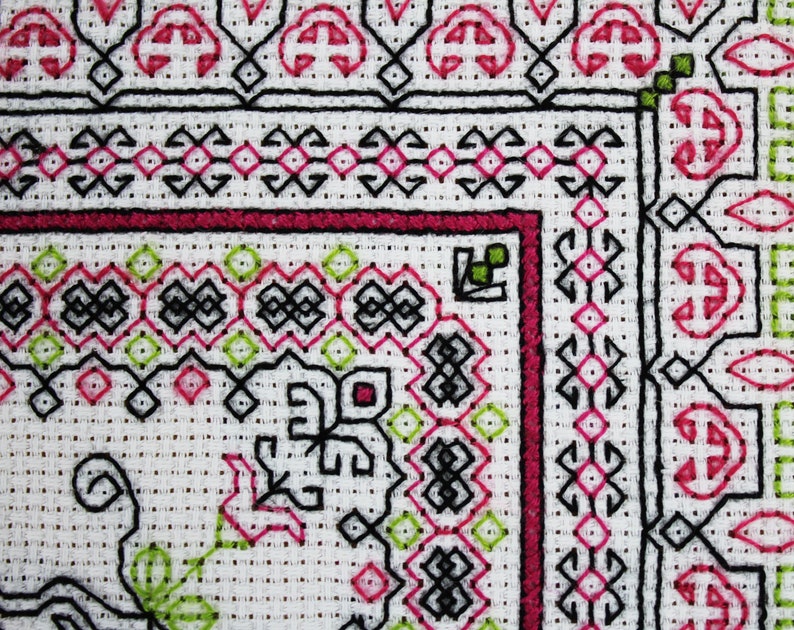 May include: Close-up of a white fabric with a black, pink, and green embroidered design. The design features geometric shapes and floral motifs.