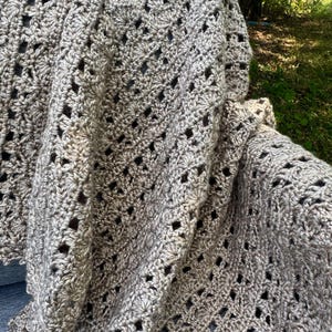 May include: A handmade, light brown crochet blanket with a textured, openwork design. The blanket is draped over a surface, showcasing the intricate stitch pattern and soft, neutral color. The blanket appears to be made of a soft yarn.