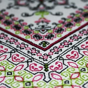May include: Close-up of a white fabric with a geometric pattern embroidered in black, pink, and green thread. The pattern features repeating shapes and lines, creating a symmetrical design.