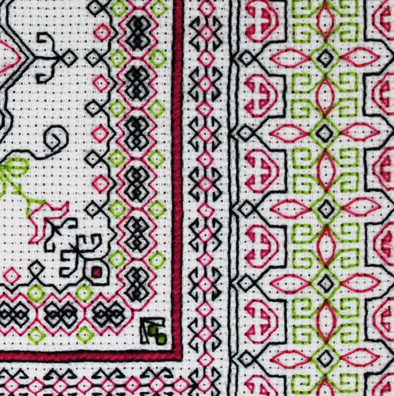 May include: Close-up of a white fabric with a black, green, and pink embroidered design. The design features geometric shapes and floral motifs.