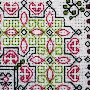 May include: Close-up of a white fabric with a black, green, and pink cross-stitch pattern. The pattern features geometric shapes and floral motifs.