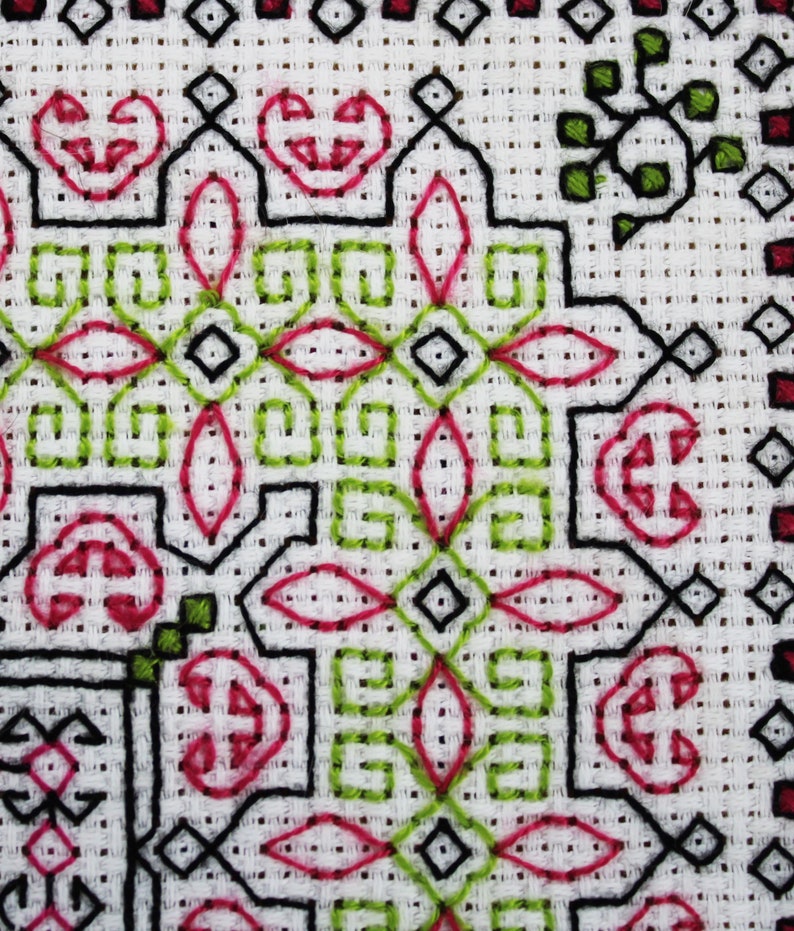 May include: Close-up of a white fabric with a black, green, and pink cross-stitch pattern. The pattern features geometric shapes and floral motifs.