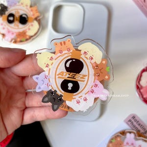 Peut inclure: Un support de téléphone en acrylique transparent avec le mot "ATEEZ" en orange, entouré de visages d'animaux de dessin animé. La poignée a un design noir et blanc au centre. D'autres supports de téléphone similaires sont visibles en arrière-plan.