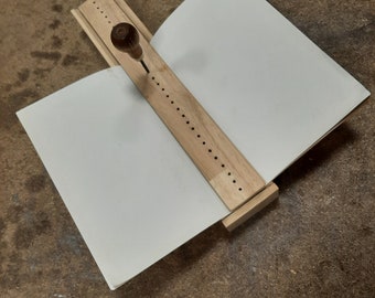 Puede incluir: Una herramienta de encuadernación de madera con una serie de agujeros y una pequeña manija de madera. Un trozo de papel blanco se coloca en la herramienta.