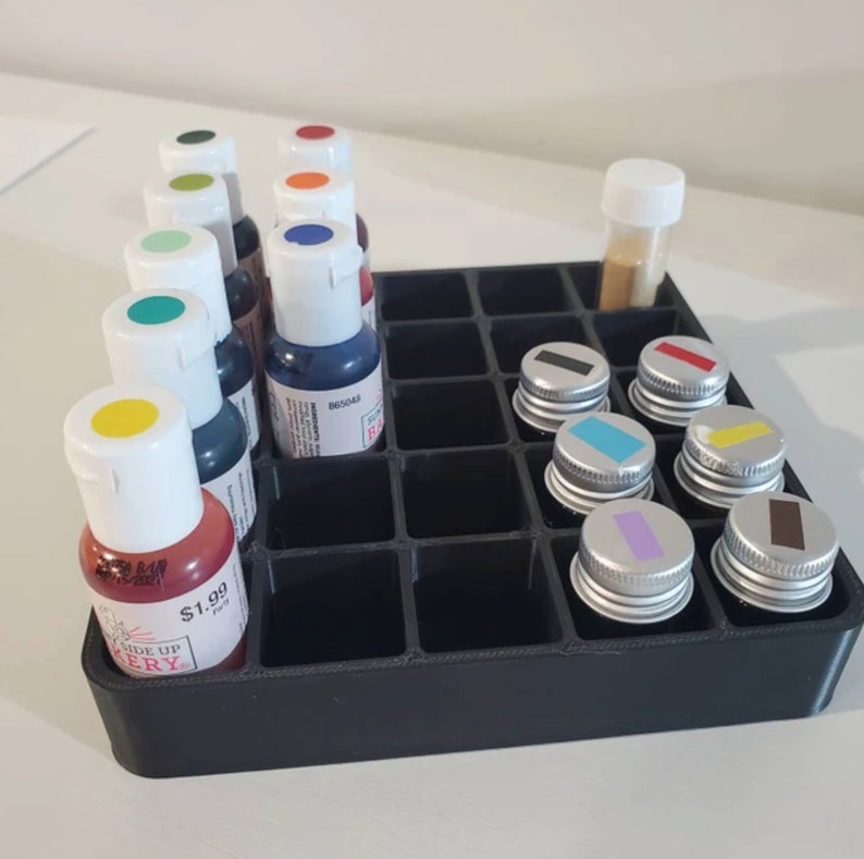 May include: A black plastic organizer with 24 compartments holds small bottles of food coloring and a small jar. The bottles are arranged in rows and have white caps. The food coloring is in various colors, including red, orange, yellow, green, blue, and purple. The organizer is on a white surface.