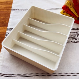May include: White plastic cutlery organizer with five compartments. The organizer is empty and sitting on a white and black patterned surface.