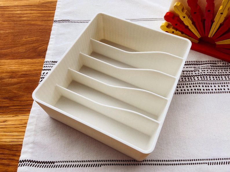 May include: White plastic cutlery organizer with five compartments. The organizer is empty and sitting on a white and black patterned surface.