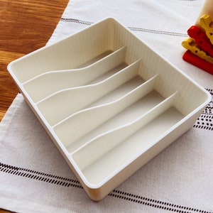 May include: White plastic cutlery organizer with five compartments. The organizer is empty and sitting on a white tablecloth with a black and white pattern.
