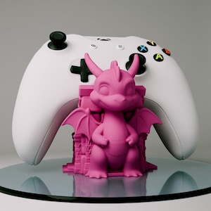 Pode incluir: Uma figura de dragão rosa com asas, sentada em uma pequena base detalhada. O dragão está segurando suas mãos na frente dele. A figura está sentada em cima de um controle Xbox branco.