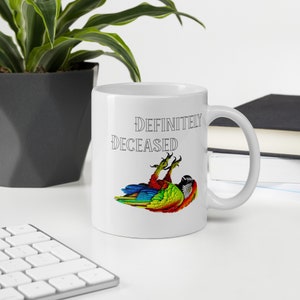 May include: White ceramic mug with the text "DEFINITELY DECEASED" in black. The mug features a colorful illustration of a parrot lying on its back. The parrot has red, blue, green, and yellow feathers. A humorous gift idea.