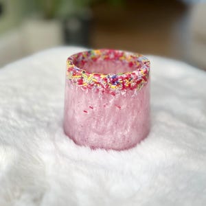 May include: A pink resin cup with a colourful sprinkle rim. The cup is sitting on a white fluffy surface.