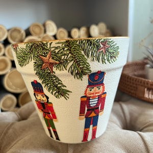 May include: White ceramic planter with a gold rim, featuring a Christmas-themed design with two nutcracker figures and pine branches with gold stars.