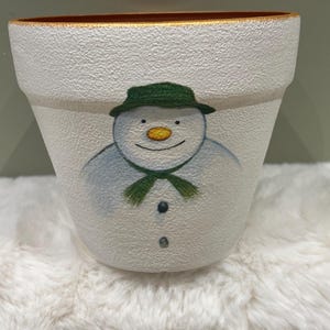 May include: A white textured plant pot featuring a hand-painted snowman wearing a green hat and scarf, with a bright orange carrot nose and two small black buttons on the chest. The rim of the pot is painted with a thin gold-coloured border.