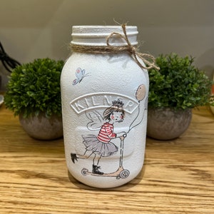 May include: White painted glass jar with a brown twine bow. The jar has a hand-painted design of a girl with wings riding a scooter with a balloon. The jar has the word "Kilner" on the front.