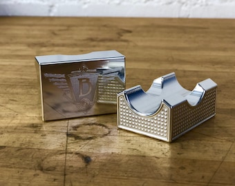 May include: Two silver metal boxes, one with a raised design and the other with a logo featuring a letter "D" inside a shield with wings.