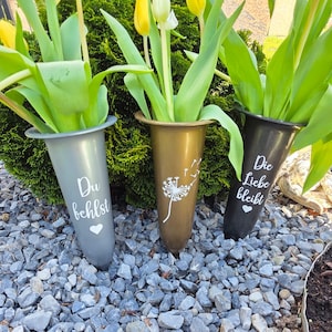 Könnte beinhalten: Drei konisch geformte Vasen in Grau, Gold und Schwarz, jede mit gelben Tulpen. Die graue Vase trägt den Text "Du behältst" und ein Herz. Die goldene Vase zeigt einen Löwenzahn. Die schwarze Vase hat "Die Liebe bleibt" und ein Herz.