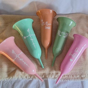 Könnte beinhalten: Fünf farbige, konisch geformte Vasen in Rosa, Grün und Orange. Jede Vase hat eine einzigartige weiße Grafik und Text. Die Vasen sind auf einem neutralen Stoff angeordnet und schaffen eine weiche, einladende Präsentation.