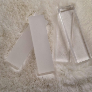 Könnte beinhalten: Drei transparente Acrylblöcke, zwei sind rechteckig und einer ist ein Trapez, auf einer weißen flauschigen Oberfläche.
