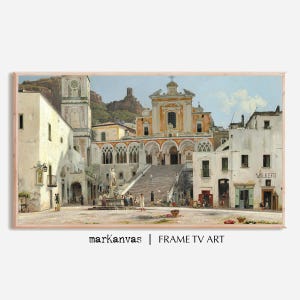 May include: Framed art print of a European town square with a church and fountain. The buildings are white with orange accents, and the sky is blue. The print is in a light brown frame. The text "markanvas | FRAME TV ART" is at the bottom.