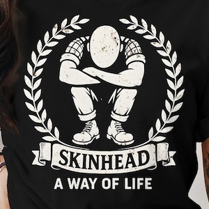 Può includere: T-shirt nera con grafica bianca di una persona seduta con le braccia incrociate, circondata da una corona d'alloro. Il testo "SKINHEAD A WAY OF LIFE" è visualizzato sotto su uno stendardo.