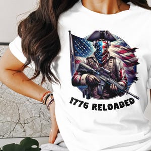 May include: White t-shirt with a graphic of a person wearing a bandana and holding a rifle. The graphic is surrounded by an American flag and the text "1776 RELOADED".