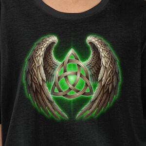 Può includere: T-shirt nero con grafica di ali d'angelo e nodo celtico. Le ali sono marroni con dettagli piumati, il nodo è verde e oro. Il disegno è centrato sulla maglietta, creando un forte impatto visivo.