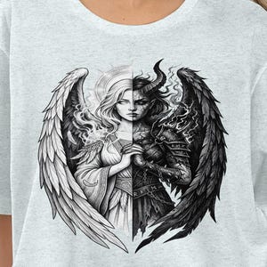 Puede incluir: Camiseta gris claro con una ilustración en blanco y negro de una mujer con alas de ángel y demonio. El diseño está dividido por la mitad, mostrando ambos lados. El rostro de la mujer está centrado, con un lado angelical y otro demoníaco.