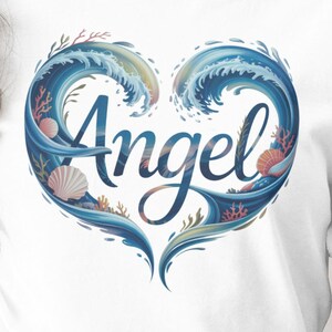 Puede incluir: Camiseta blanca con un diseño en forma de corazón con olas del océano, conchas marinas y coral. La palabra "Angel" está escrita en una fuente cursiva dentro del corazón. El diseño es en tonos azules y coral.