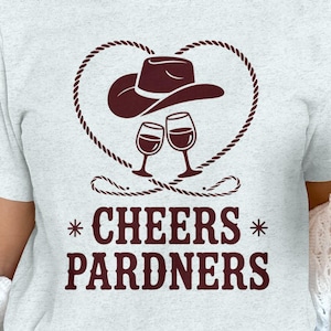 May include: Pale grey t-shirt featuring a brown graphic of a cowboy hat, wine glasses, and the words "Cheers Pardners." A Santa-themed mug is held in the hand. The shirt design is centered on the chest.