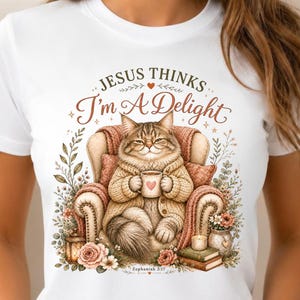 May include: White t-shirt featuring a watercolor illustration of a cat in a cozy armchair, holding a mug with a heart. The text "Jesus Thinks I'm A Delight" is above the cat. Floral and book accents complete the design.
