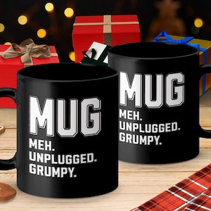 May include: Two black ceramic mugs with the word "MUG" in large white letters, and the text "MEH. UNPLUGGED. GRUMPY." The mugs are on a wooden surface with Christmas gifts in the background.