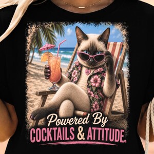 May include: A black t-shirt featuring a graphic of a Siamese cat wearing pink sunglasses and a floral shirt, sitting in a beach chair with a cocktail. The text below reads, Powered By COCKTAILS & ATTITUDE. The design includes a beach and ocean scene.