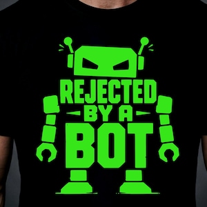 May include: A black t-shirt featuring a bright neon green graphic of a robot. The text REJECTED BY A BOT is printed in large, bold, block letters across the center of the robot's torso, with the robot's head and limbs framing the text.