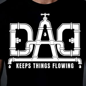 Puede incluir: Camiseta negra con un diseño gráfico blanco. El diseño forma la palabra "DAD" con tuberías de fontanería y un grifo. Debajo, el texto "KEEPS THINGS FLOWING" en blanco.