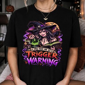 Può includere: Una t-shirt nera con la grafica di una strega con un cappello viola, che mescola una pozione verde in un calderone tenendo in mano un libro aperto. Il testo recita "YOU HAD ME AT TRIGGER WARNING" in lettere arancioni e viola con uno sfondo lunare.