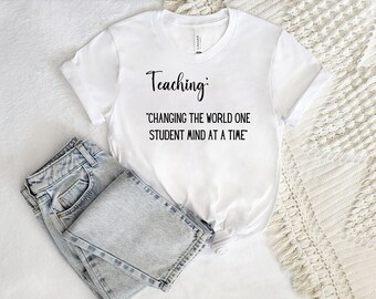 May include: White t-shirt with the text "Teaching: Changing the world one student mind at a time" printed on it. The shirt is lying on a white surface with a pair of light blue jeans and a white crocheted blanket.