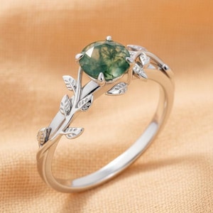 May include: A silver ring with a round, green moss agate gemstone. The ring features a delicate leaf design on the band, with small leaves accenting the gemstone setting. The ring is set against a peach-colored fabric.
