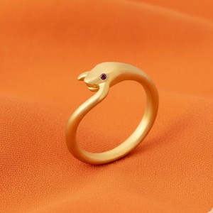 Può includere: Un anello color oro a forma di delfino, con una piccola gemma rossa e rotonda per l'occhio. L'anello è posto su uno sfondo arancione.