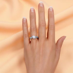 May include: A silver ring with a round, white gemstone is displayed on a hand. The ring features a floral design with delicate leaves and branches. The background is a soft peach color.