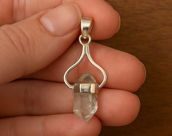 May include: A clear, pointed quartz crystal pendant set in a polished silver-toned metal cap with a decorative, arched bail. The crystal has a natural, multifaceted hexagonal shape and is suspended from a smooth, curved silver-toned metal frame.